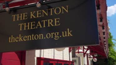 A black sign for The Kenton Theatre displays the website thekenton.org.uk in gold lettering, mounted on a building with red trim and white brick.