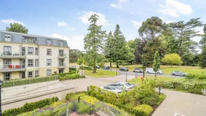 View of a modern apartment building with balconies, landscaped gardens, parked cars, and a background of large trees under a partly cloudy sky.