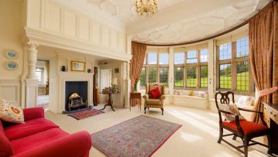 Elegant living room with large bay windows, red sofas, an ornate rug, and a fireplace. A chandelier hangs from a detailed ceiling, and the room overlooks a garden.