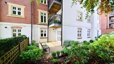 Ground-floor apartment with red and white brick exterior, a balcony, and a small garden area with a bench and plants.