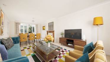 Spacious living room with a blue sofa, yellow armchairs, a large TV, and a dining table with yellow chairs. A colorful rug covers the floor, and a bouquet of flowers is on the glass coffee table.