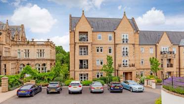 Parking lot with six cars in front of a modern stone apartment building, with an older stone building to the left and greenery around.