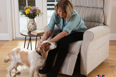 A woman sitting on a recliner gently pets a brown and white dog. A vase with flowers and a book are on the table nearby. A logo in the corner indicates the chair is handmade in the UK.