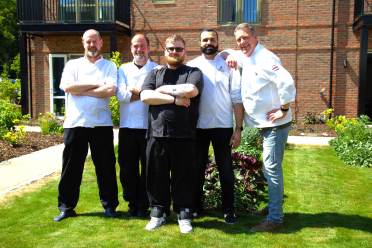 Five men in chef uniforms stand together on a sunny lawn in front of a brick building, posing and smiling at the camera.