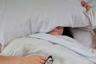 A person lying in bed partially covered by a pillow, holding a pair of glasses in one hand.