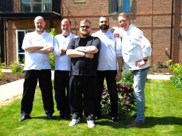 Five men in chef uniforms stand together on a sunny lawn in front of a brick building, posing and smiling at the camera.
