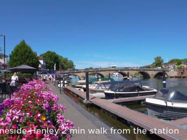 A riverside walkway in Henley with boats docked, colorful flowers in the foreground, and a stone bridge in the background; text reads "Thameside Henley 2 min walk from the station.