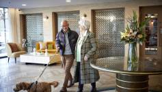 An older couple walks a small dog through a hotel lobby, decorated with a vase of flowers on a round table and patterned glass panels in the background.